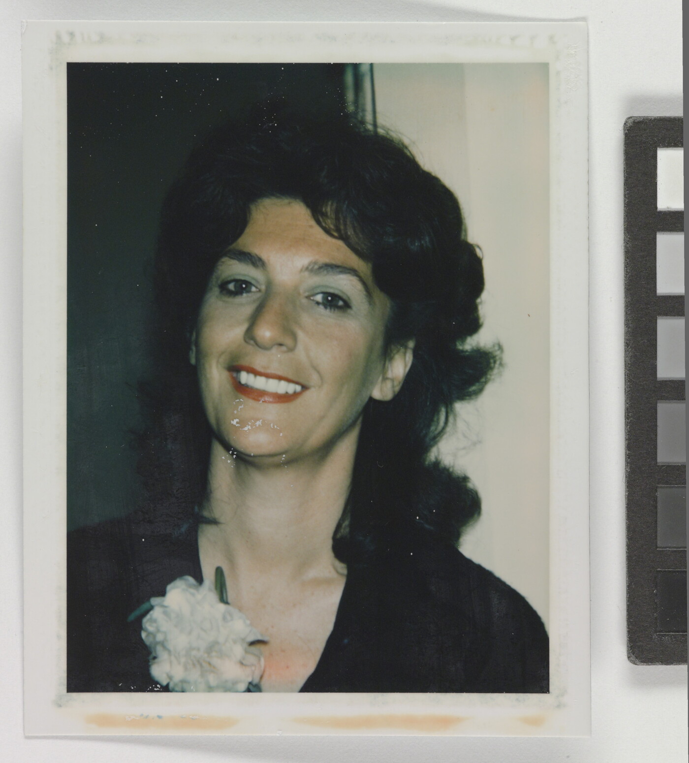 An informal color photograph of a smiling White woman from the shoulders up with dark, curly hair, red lipstick, and a white carnation on her black shirt.