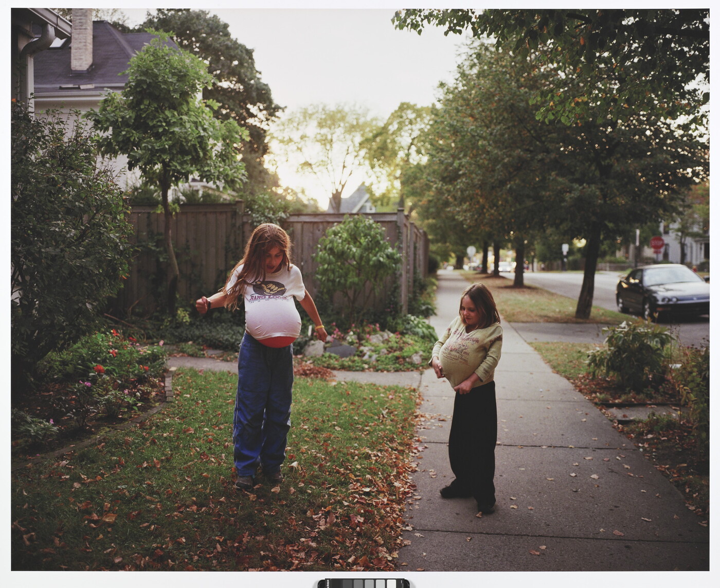 A color photograph of two young White girls playing in a front yard with balloons under their shirts mimicking pregnancy.