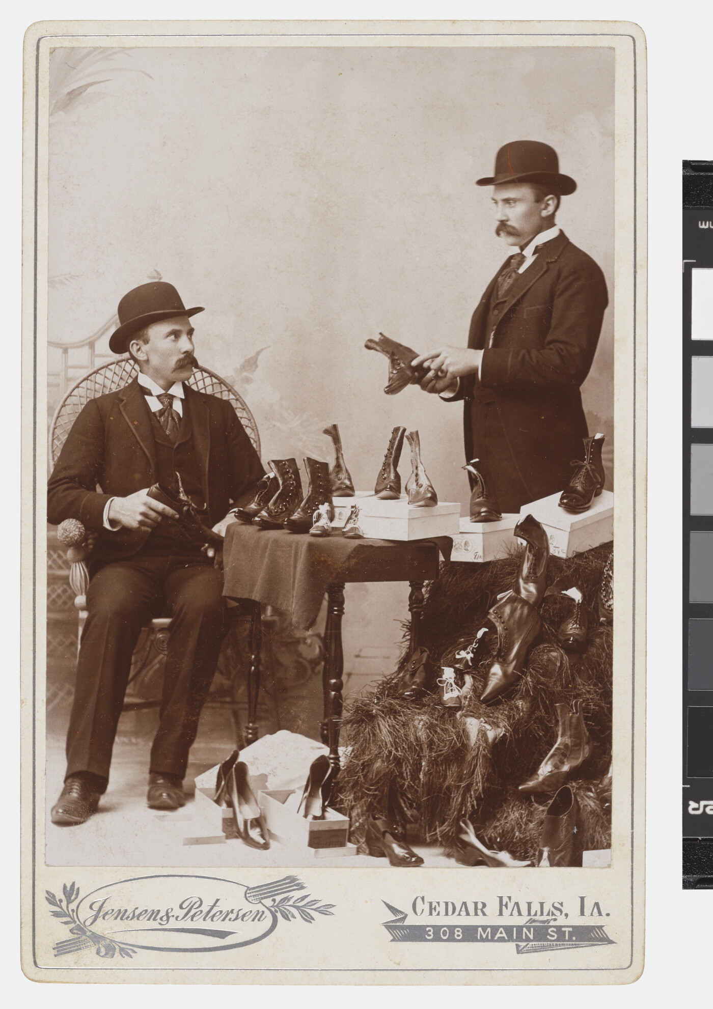 A black-and-white studio photograph of a seated White man in a bowler hat looking at shoes as the same man standing appears to show him shoes.