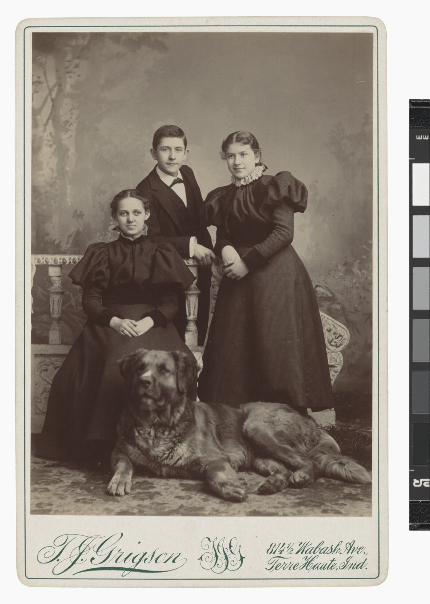 A black-and-white studio portrait photograph of three young adults, two White women and one White man, posing behind a large dog.