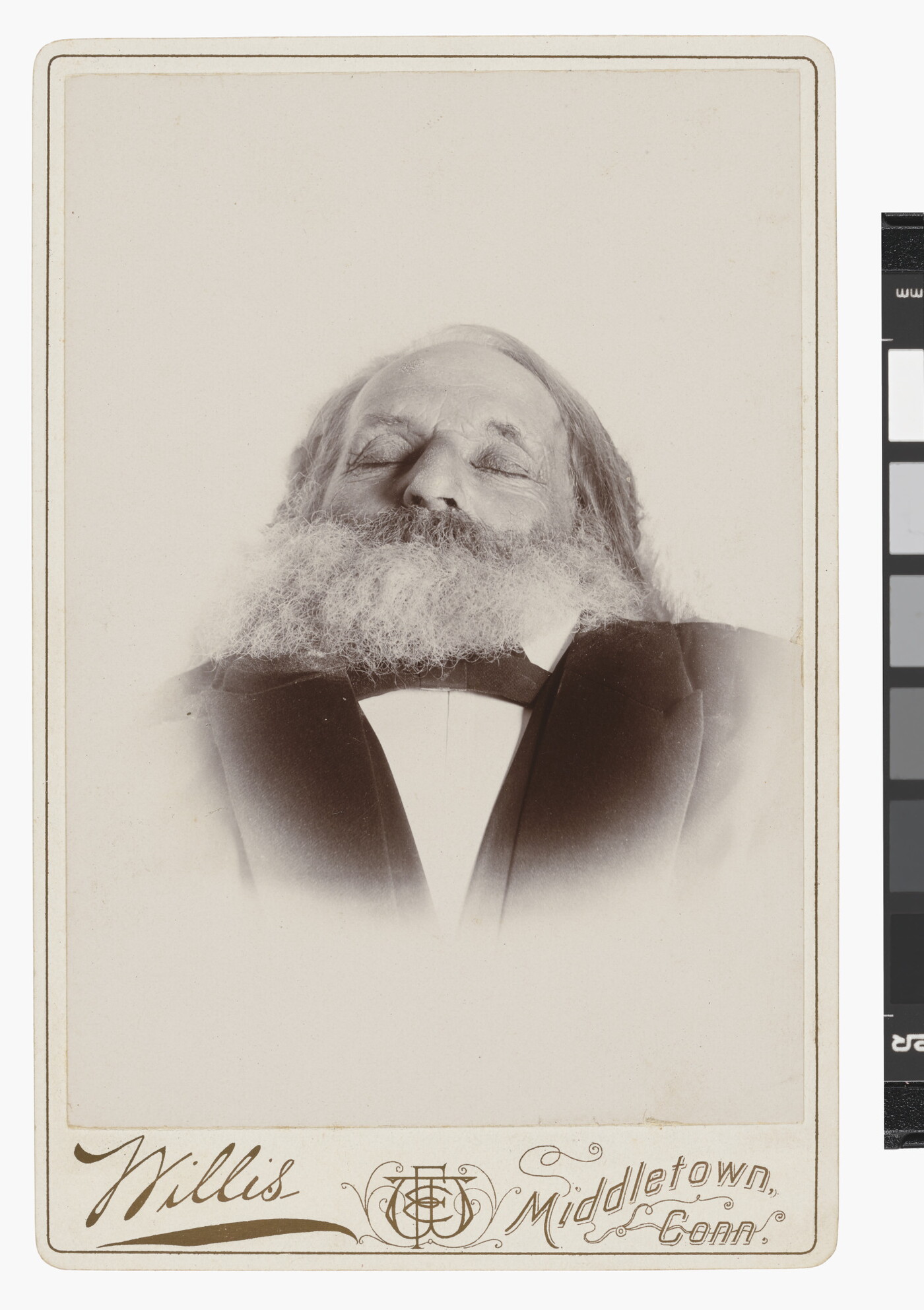 A black-and-white photograph of a deceased White man, from the shoulders up, with a full beard and eyes closed wearing a suit.