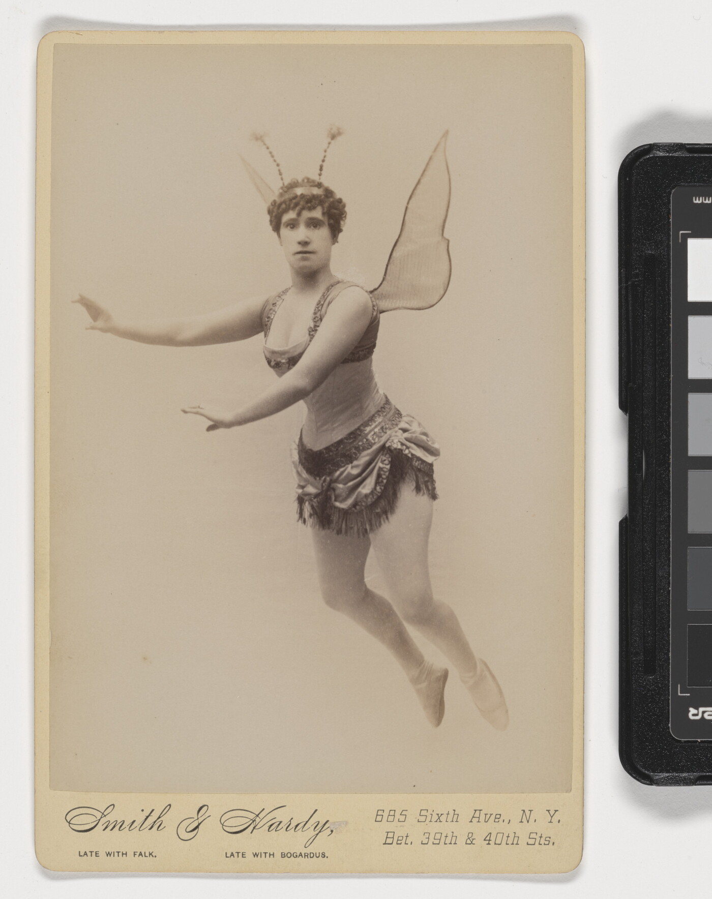 A sepia-toned studio photograph of a White woman dressed as a fairy, including wings and antennae, suspended in the air against a white background.