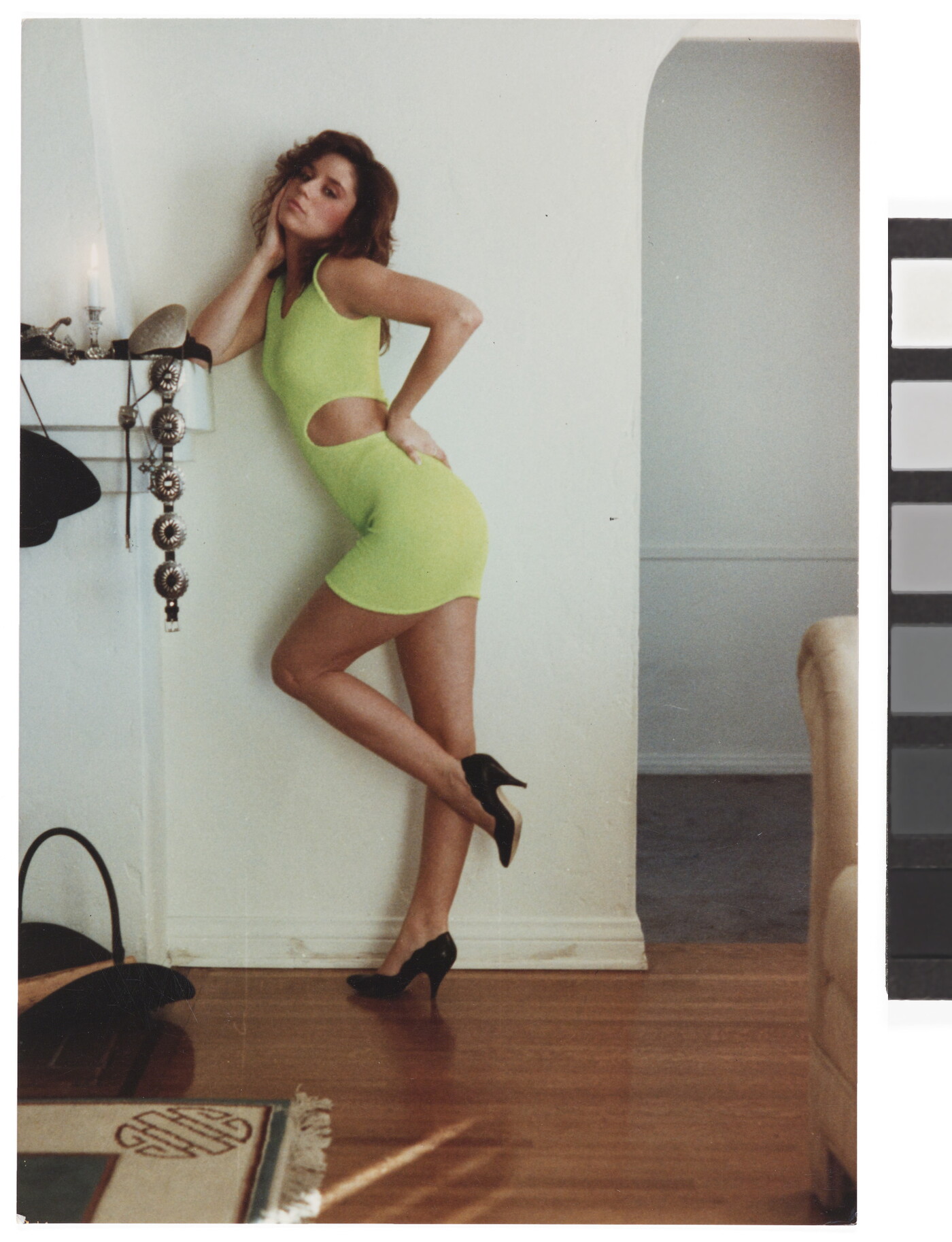 A color photograph of a White woman in a bright green dress and black heels, one hand on her hip the other elbow on a fireplace mantel, striking a pose for the camera.