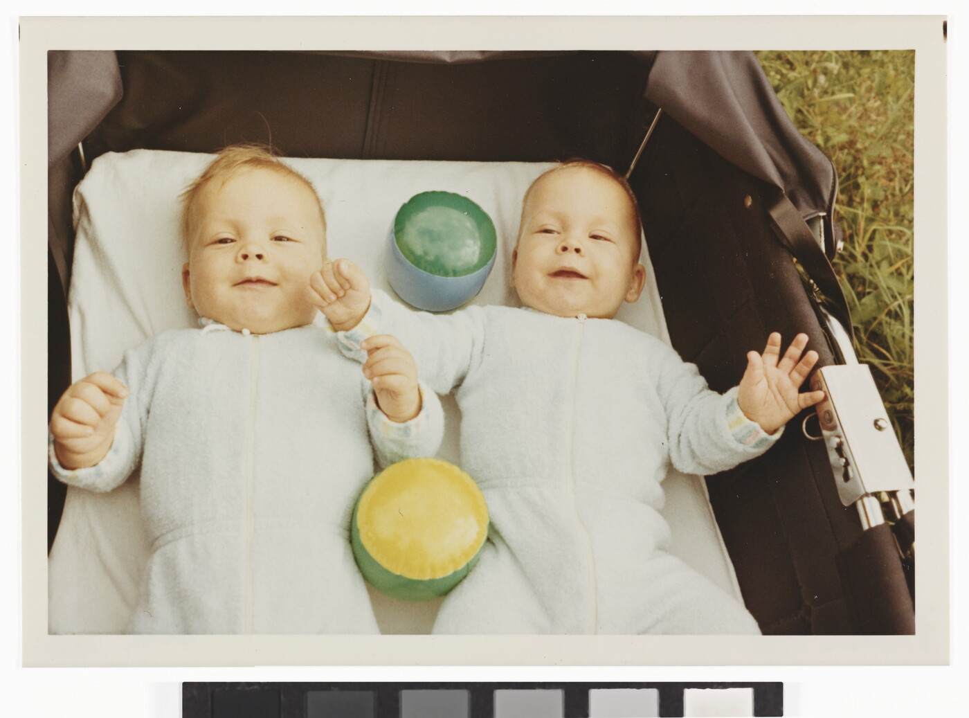 A color photograph of two White babies smiling and lying on their backs with toys in a baby carriage that is sitting in grass.