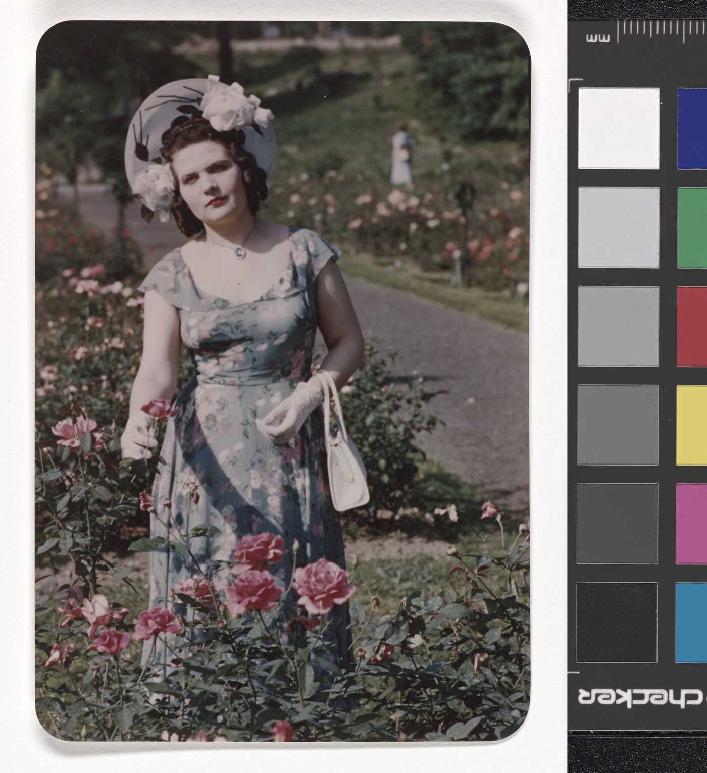 A color photograph of a White woman wearing a sunhat, floral dress, and white gloves standing in a rose garden.