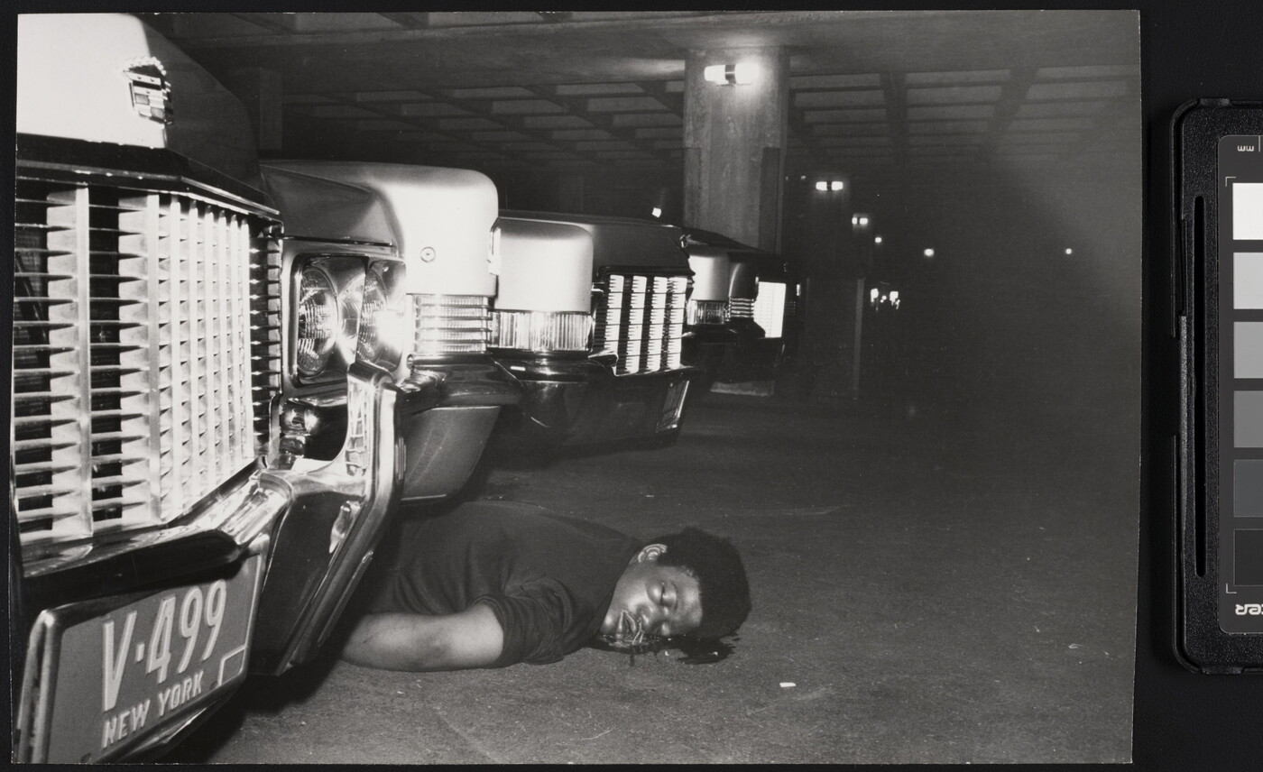 A black-and-white photograph of the body of a Black person on their stomach with a blood stain around their head between cars in a parking garage.