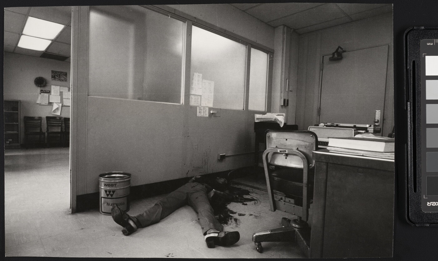 A black-and-white photograph of an office with a bloody body on the floor, feet toward the camera, the top part of the body obscured by dark stains on it, the floor, and the wall.