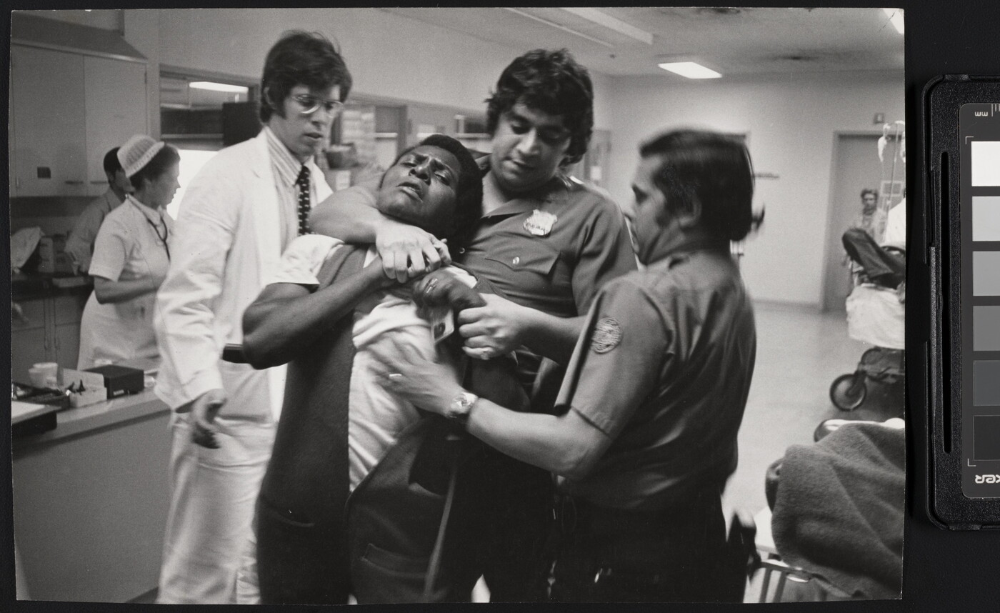 A black-and-white photograph of a Black man being restrained and choked by two police officers with medium to dark skin in a hospital setting.