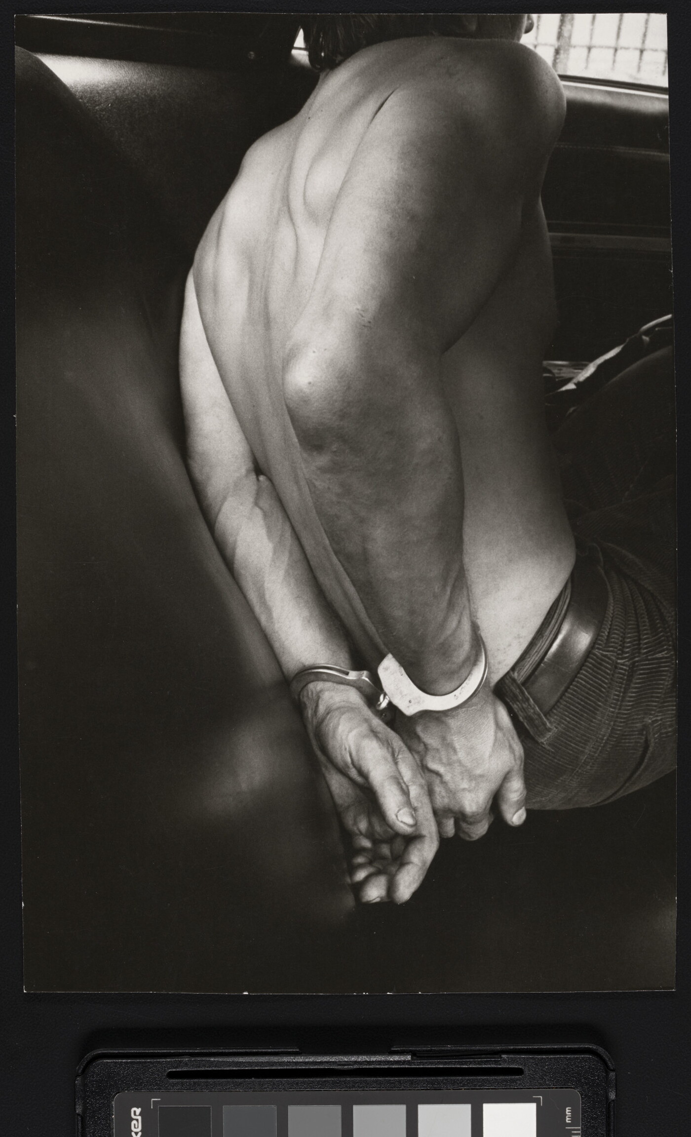 A black-and-white photograph of the bare back and handcuffed hands of a White man in the back seat of a police car.