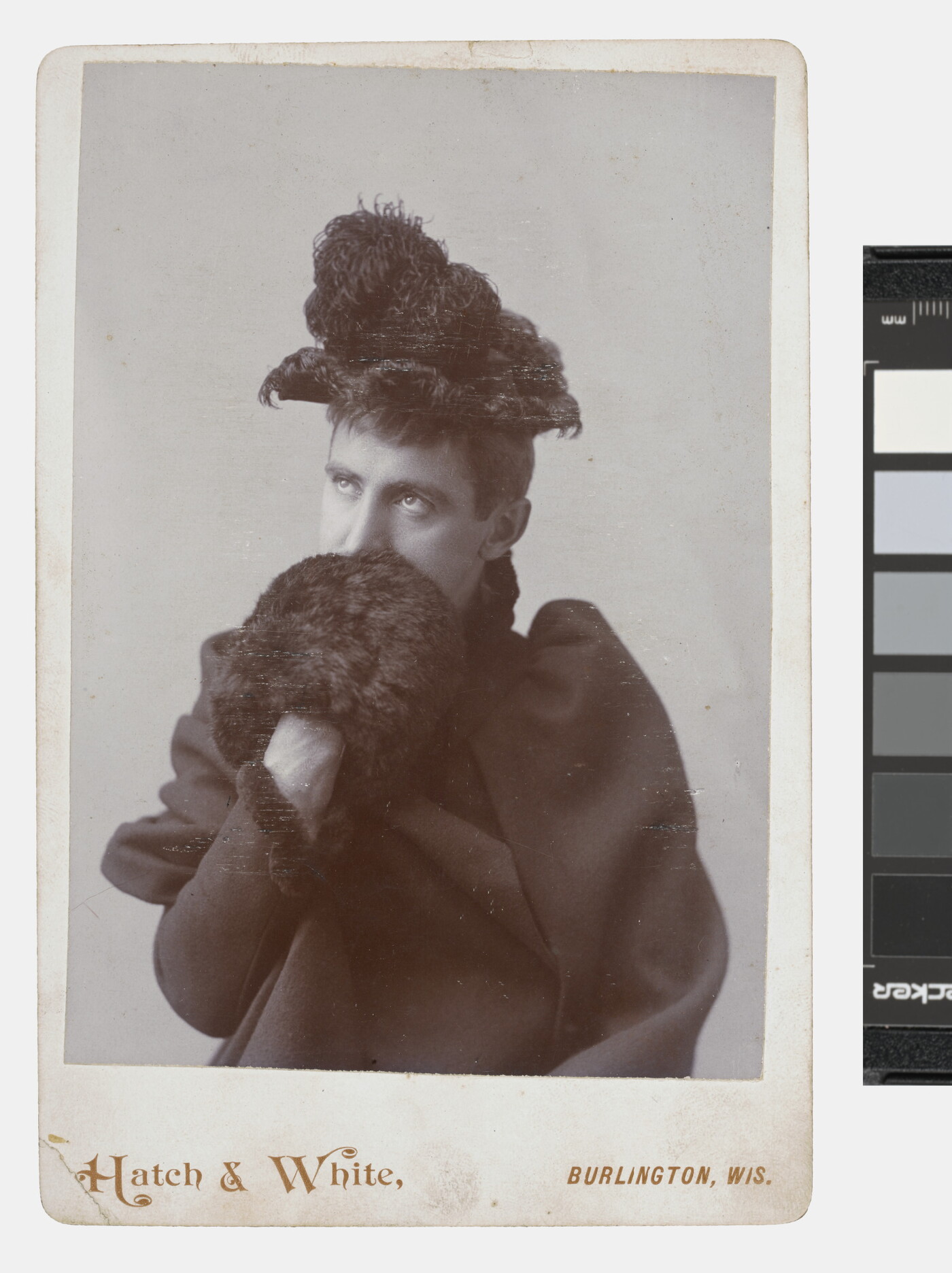 A black-and-white studio portrait photograph of a White person wearing a feather-trimmed hat, dark coat, and a fur muff that they are holding up in front of their mouth.