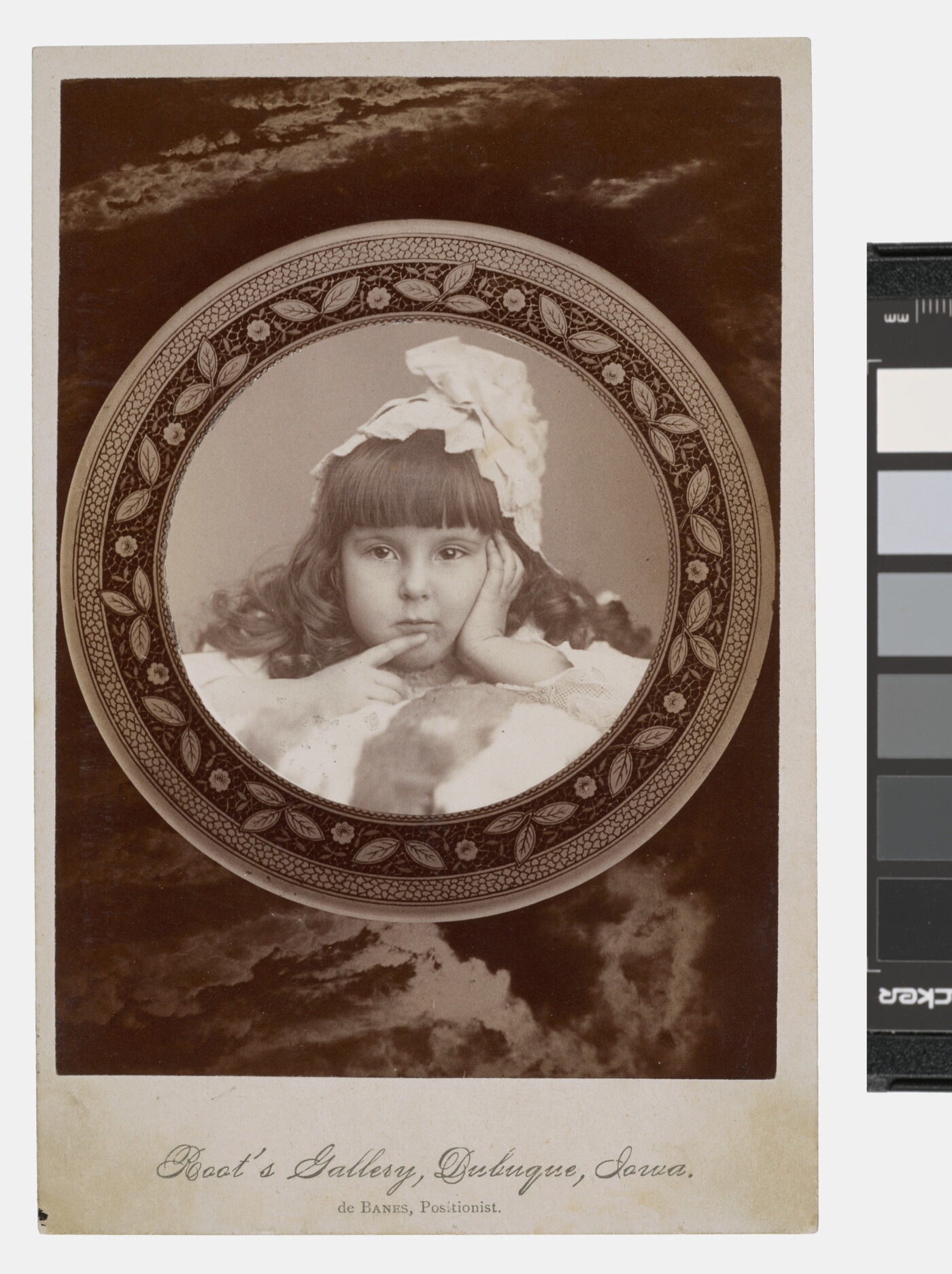 A black-and-white studio photograph of a young White girl with curled dark hair, bangs, and a massive bow on her head holding one finger to her mouth.