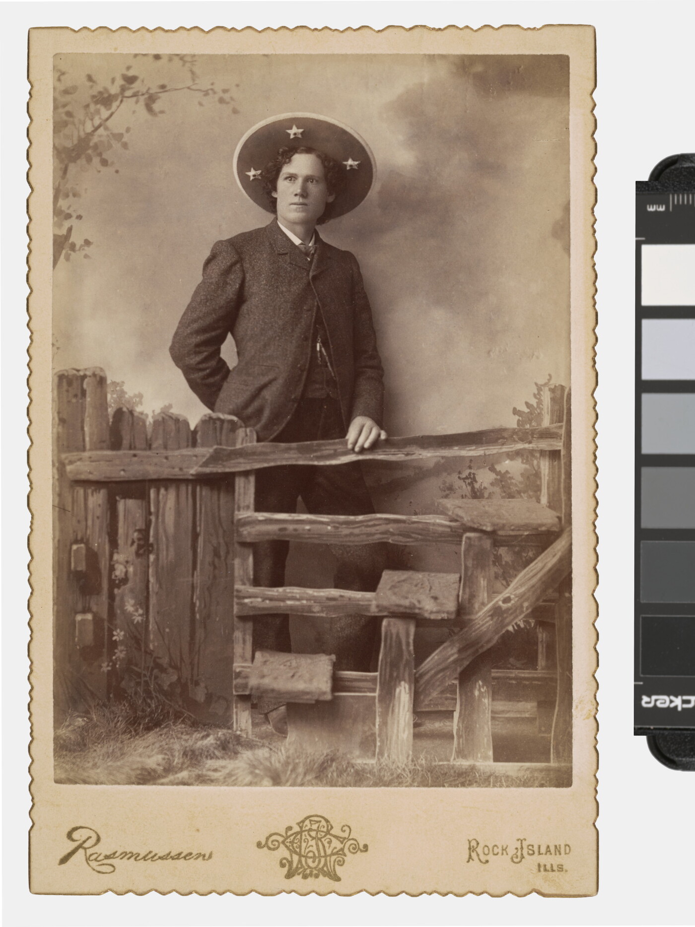 A sepia-toned photograph of a White man wearing a large sombrero-like hat standing behind a split-rail fence that is part of a studio set.