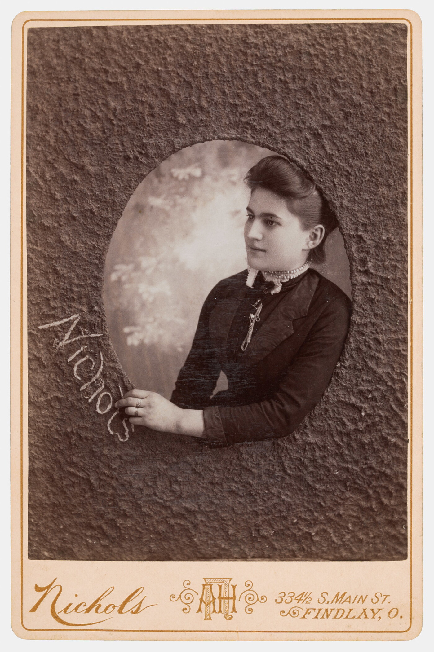 A sepia-toned studio photograph of a White woman who appears to be reaching through a textured oval window to write the word, &quot;Nichols.&quot;