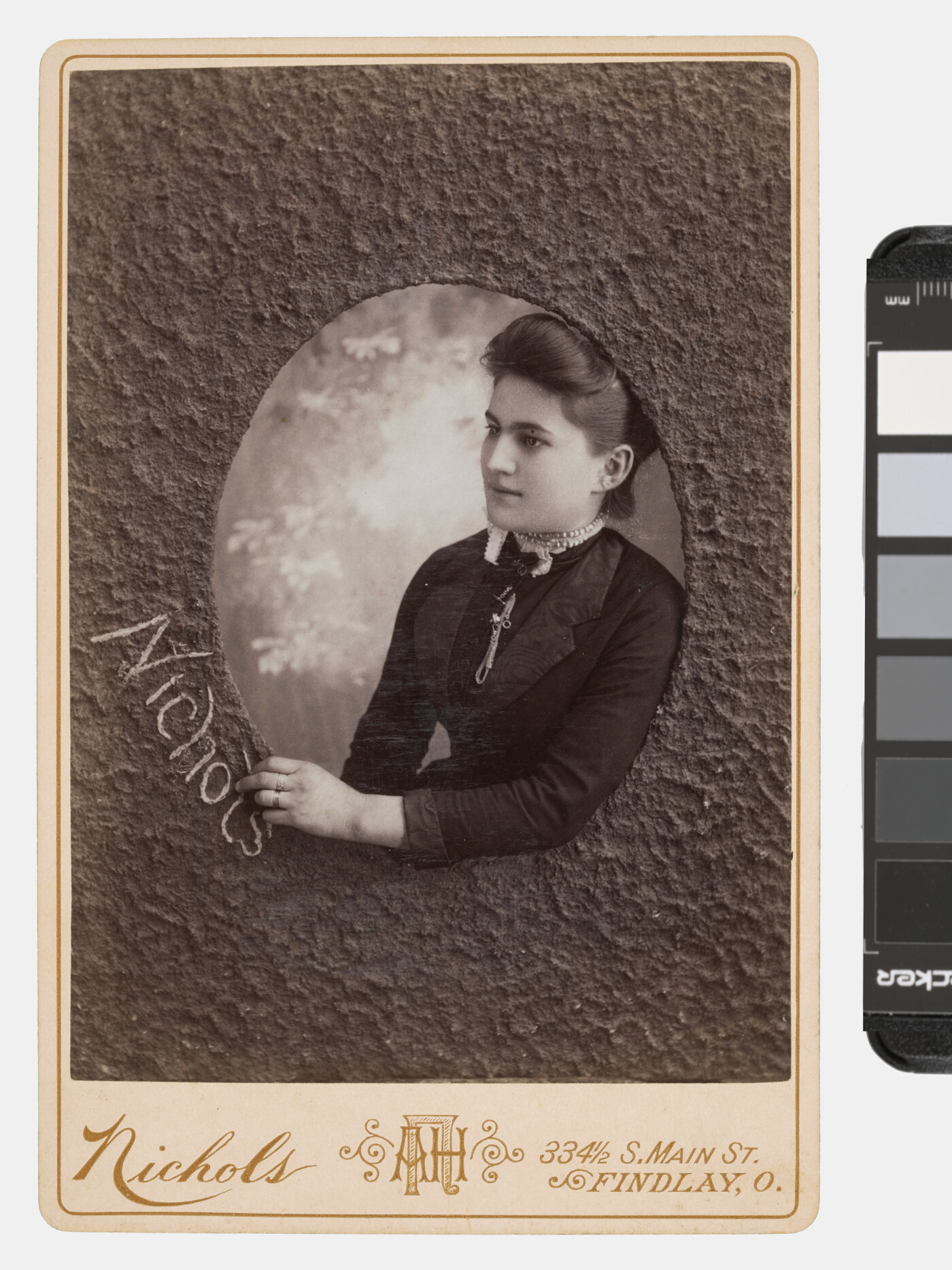 A sepia-toned studio photograph of a White woman who appears to be reaching through a textured oval window to write the word, &quot;Nichols.&quot;