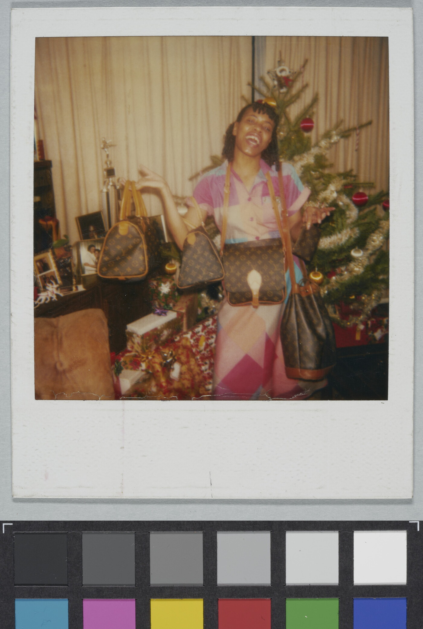 A color Polaroid photograph of a Black woman gleefully showing off several designer purses in front of a Christmas tree.