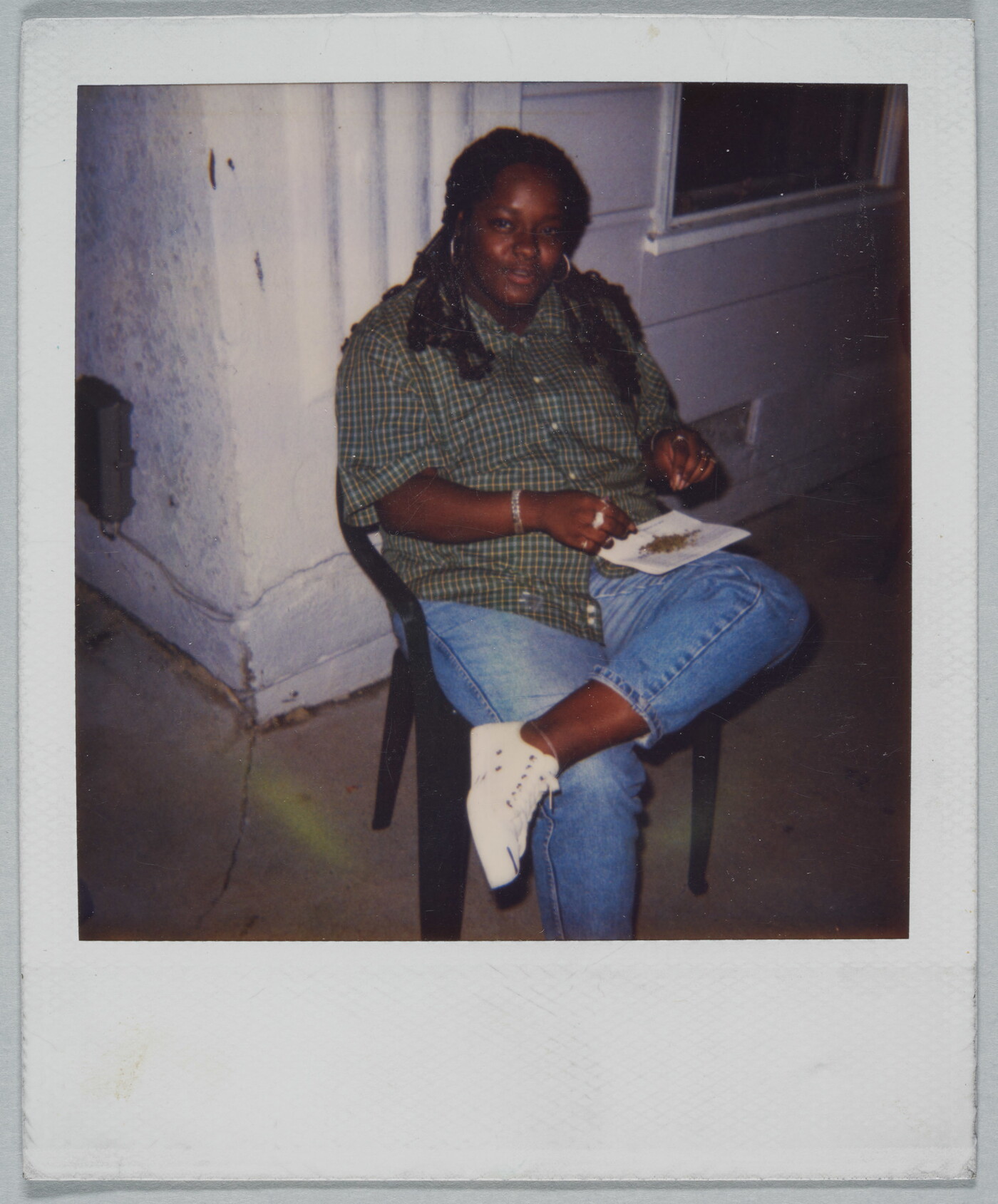 A color Polaroid photograph of a Black woman seated on a plastic chair balancing a piece of paper with bits of something on it on her crossed leg.