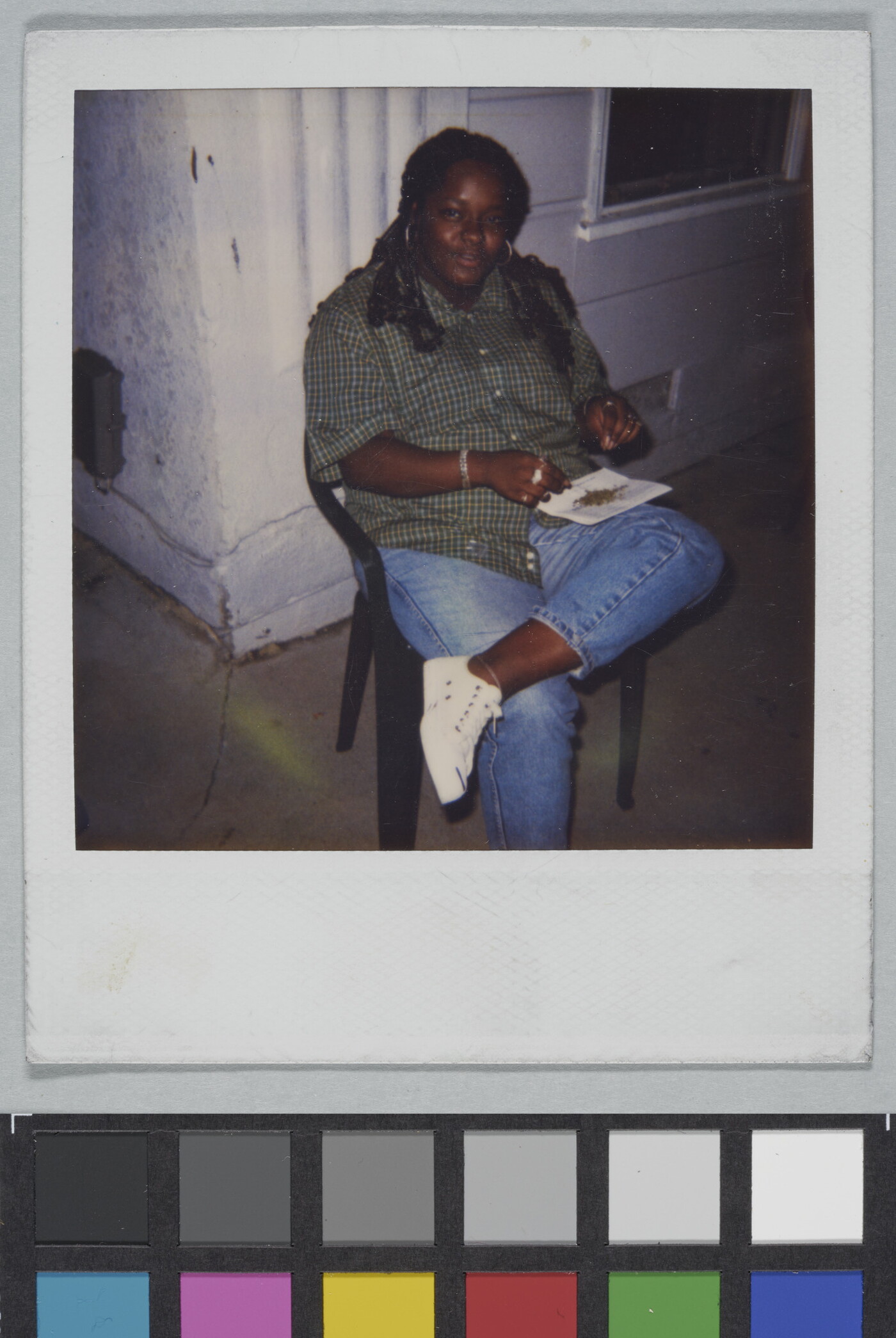A color Polaroid photograph of a Black woman seated on a plastic chair balancing a piece of paper with bits of something on it on her crossed leg.