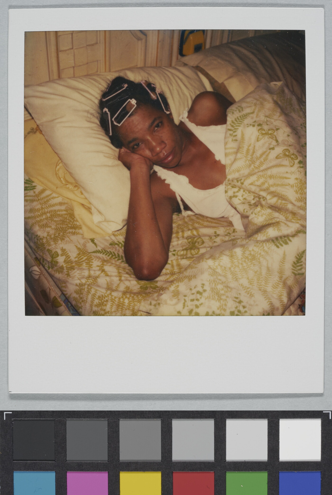 A color Polaroid photograph of Black woman, curlers in her hair, in bed with her head propped up on her hand.