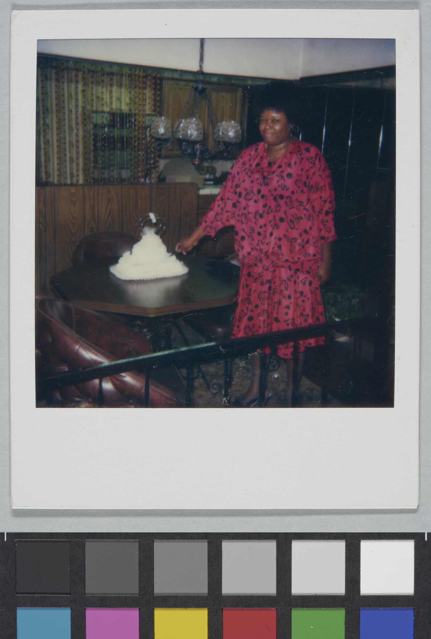 A color Polaroid photograph of a smiling Black woman in a pink and black dress slicing a tiered white cake on a dining table.