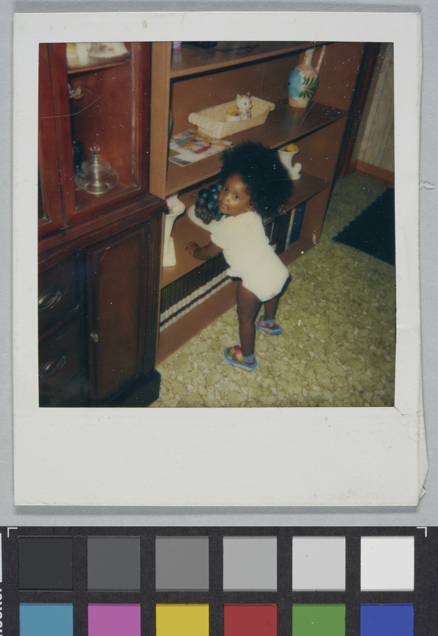 A color Polaroid photograph of Black toddler in a shirt, diaper, and sandals with big, natural hair propping themselves up on a bookshelf.