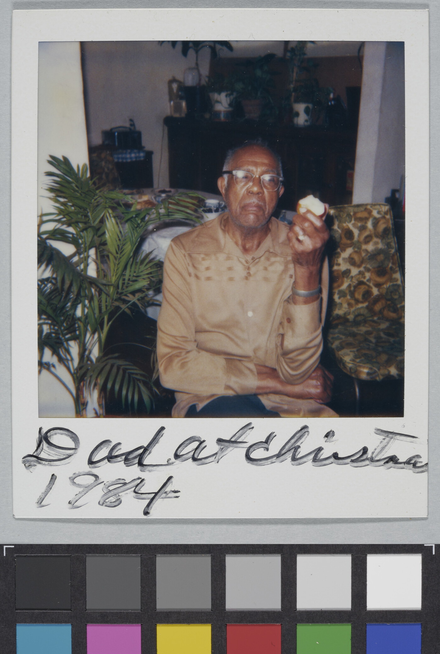 A color Polaroid photograph of a seated, middle-aged Black man in glasses and a light brown shirt holding an apple in one hand; handwritten text at bottom reads: &quot;Dad at Christma[s] 1984.&quot;
