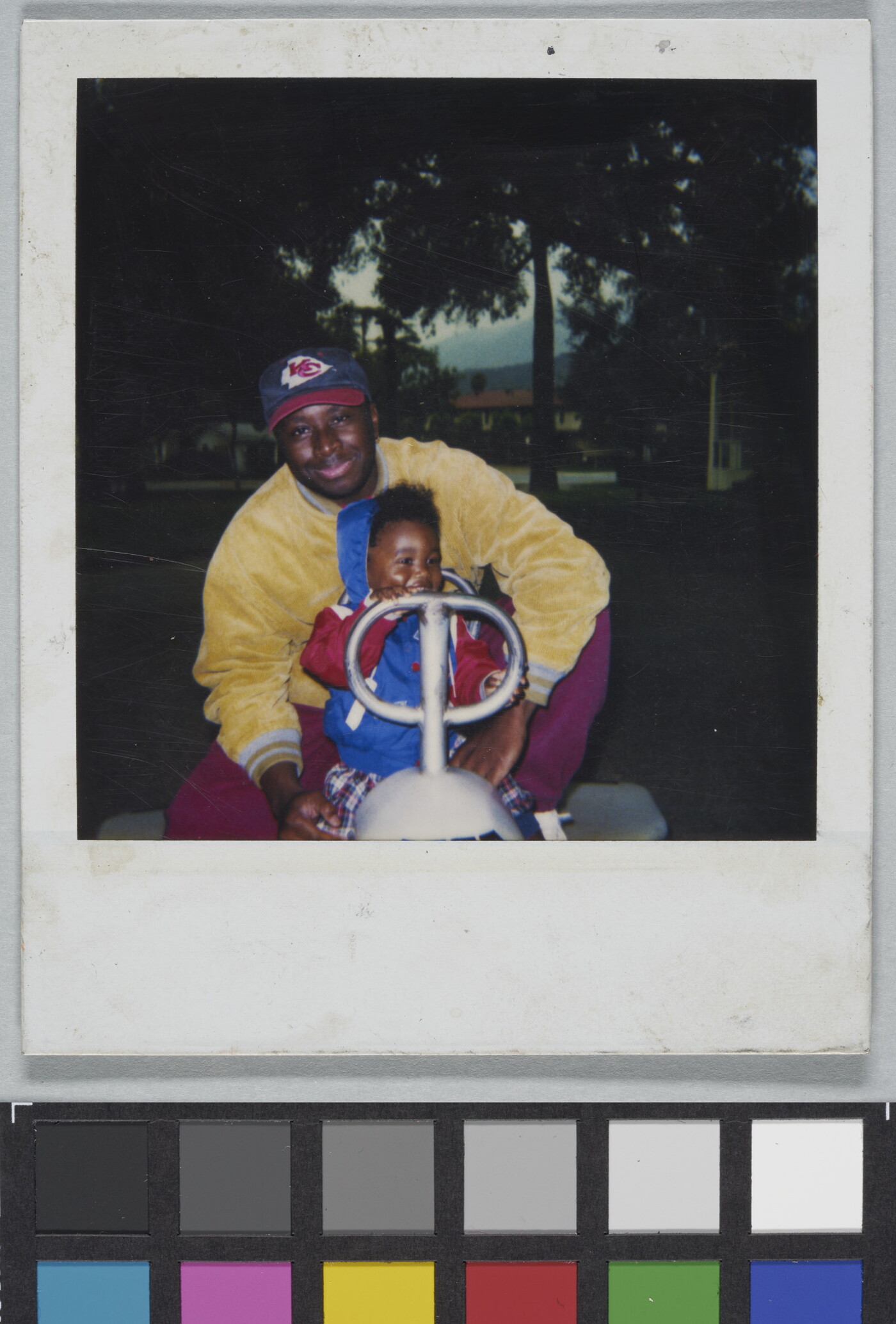 A color Polaroid photograph of a Black man and toddler seated behind the handles of an outdoor play toy in a park.