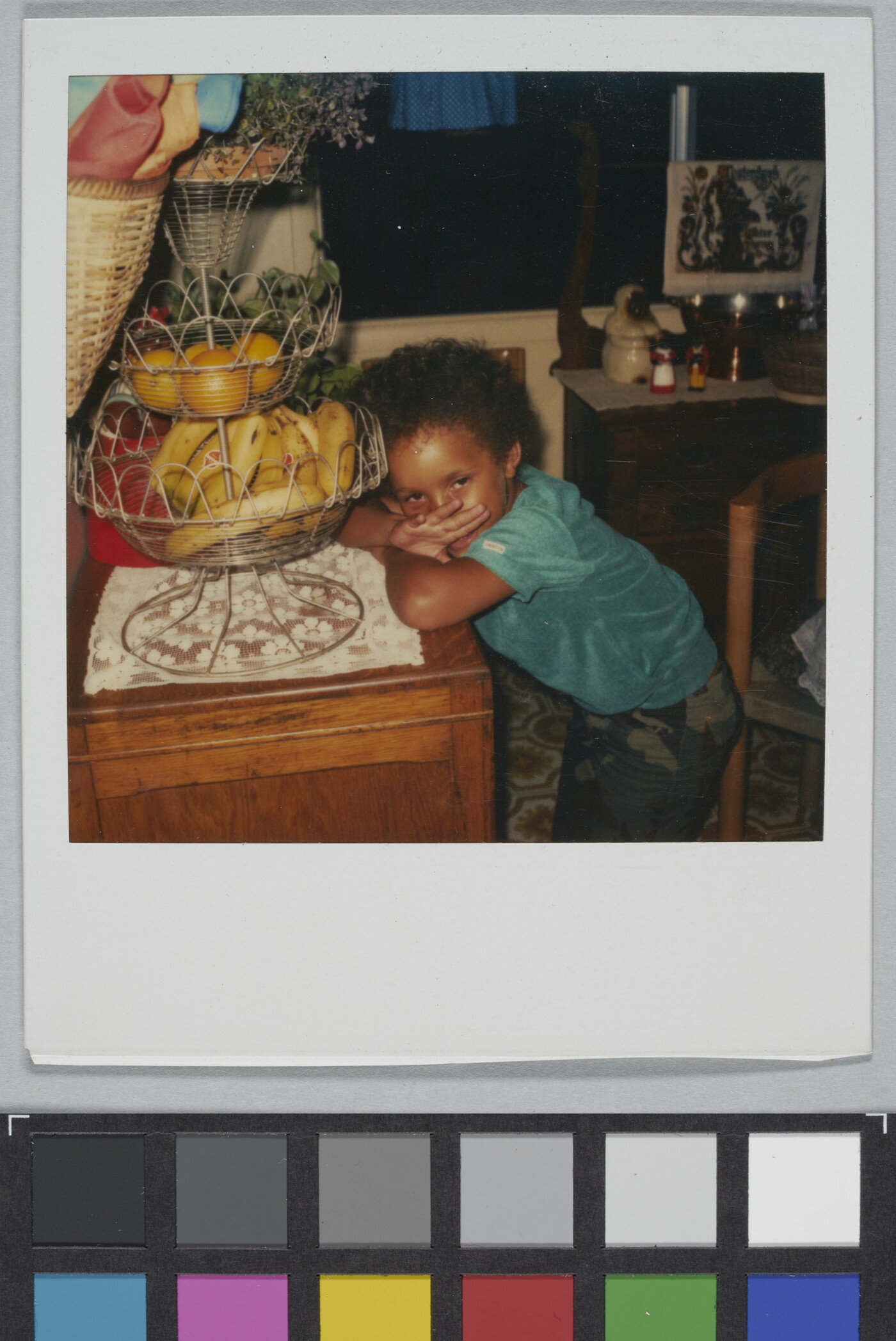A color Polaroid photograph of a Black child leaning on a table that has a tiered fruit holder.