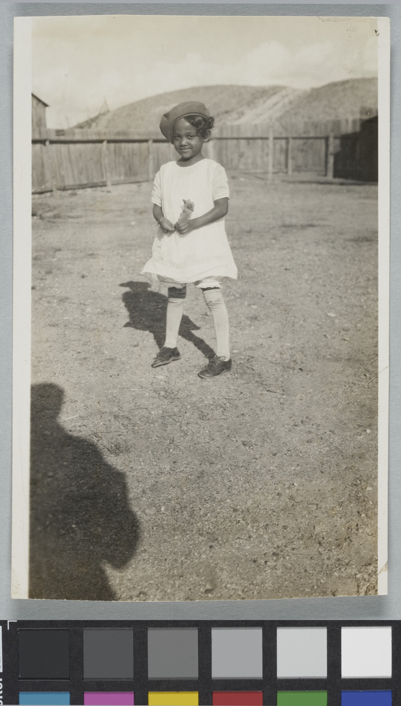 A black-and-white snapshot of a Black girl in a white dress and knee socks standing in a fenced yard.