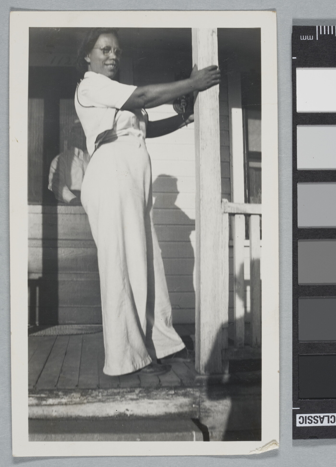 A black-and-white snapshot of a Black woman in white shirt and pants standing on a porch holding onto a column.