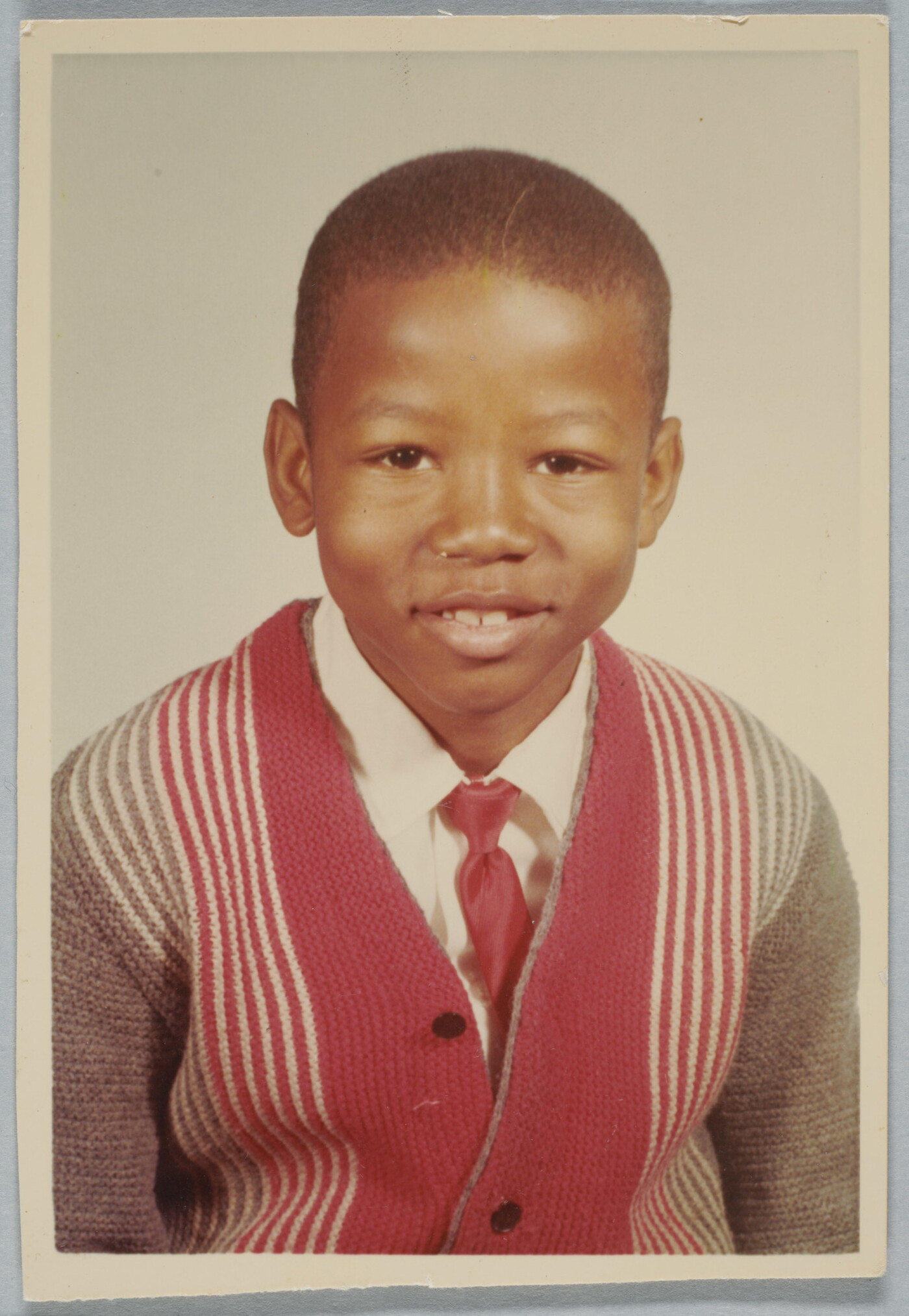 A color school photograph of a young, smiling Black boy wearing a white collared shirt and red tie under a striped cardigan.