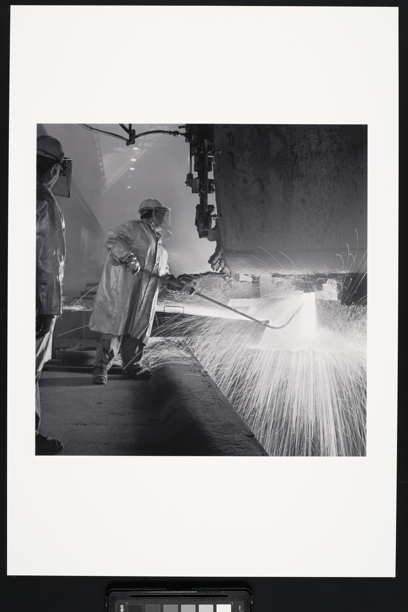 A black-and-white photograph of an industrial worker, wearing safety helmet, coat, and gloves holding a rod under a machine emitting sparks.