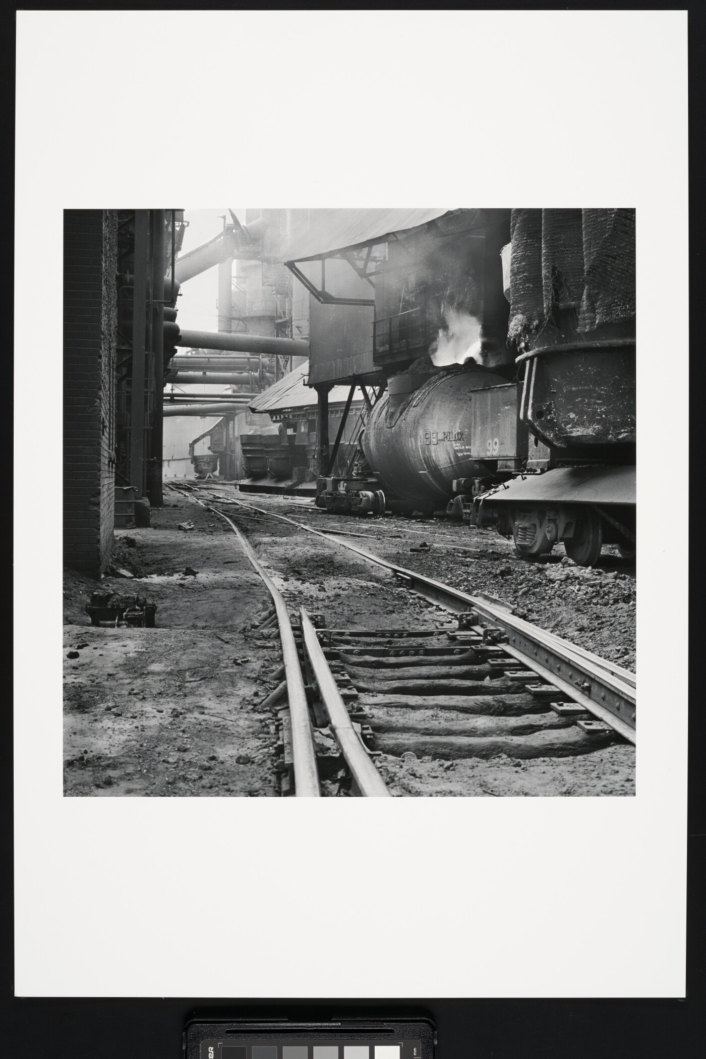 A black-and-white photograph of railroad track  between industrial buildings and  machinery, some emitting steam or smoke.