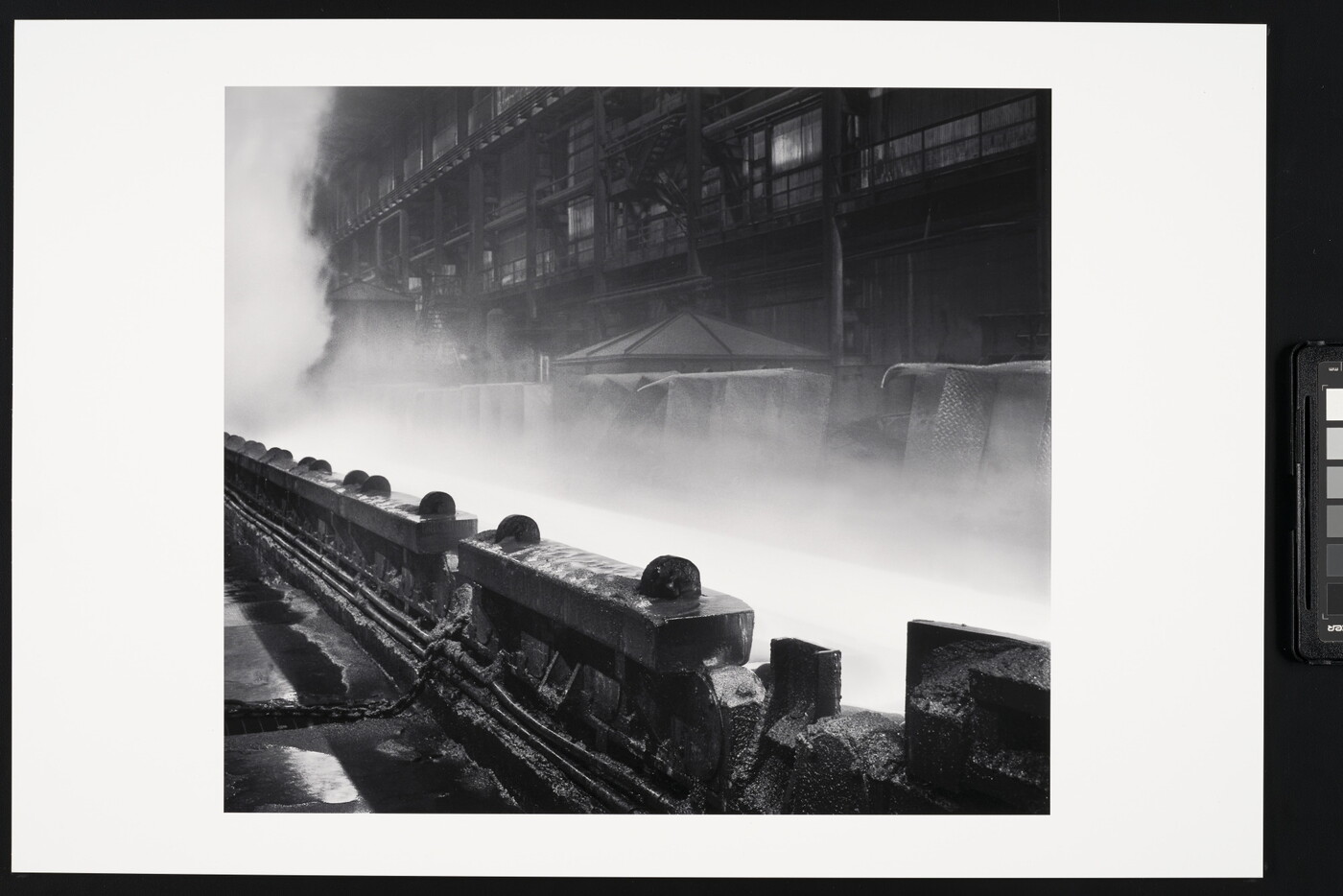 A black-and-white photograph of industrial machinery in front of steam or smoke rising near a multi-story industrial building.
