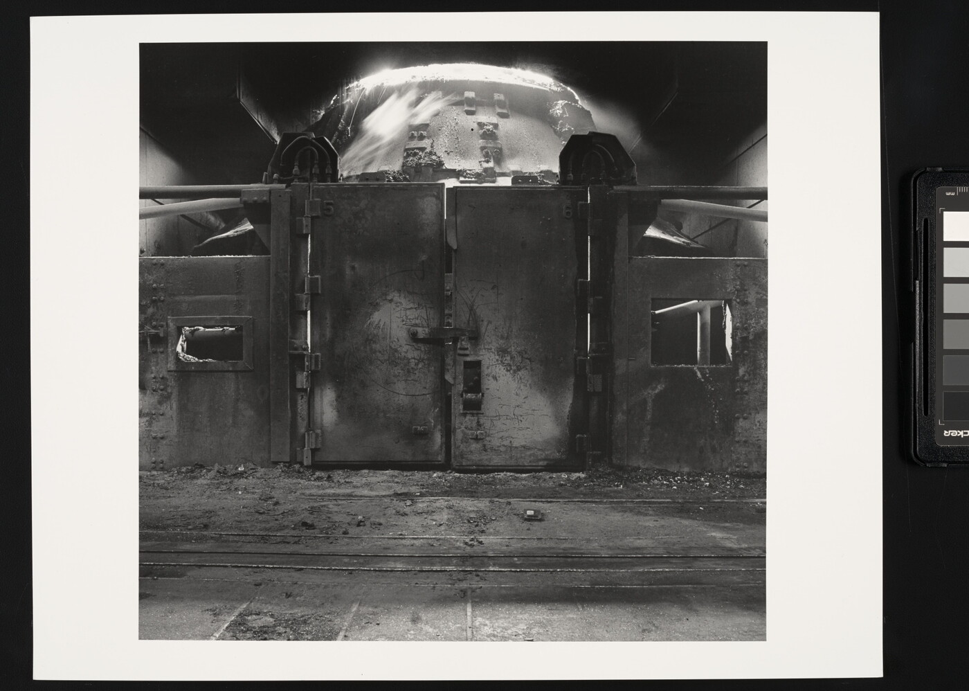 A black-and-white photograph of a metal wall with large doors behind which is an industrial furnace.