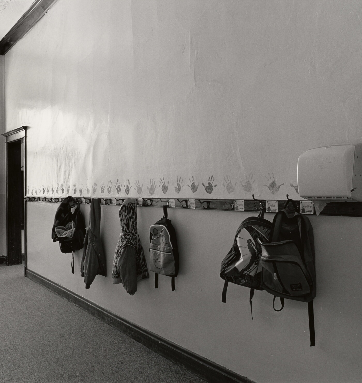 A black-and-white photograph of a hallway with kids&#039; backpacks and jackets hanging on hooks.