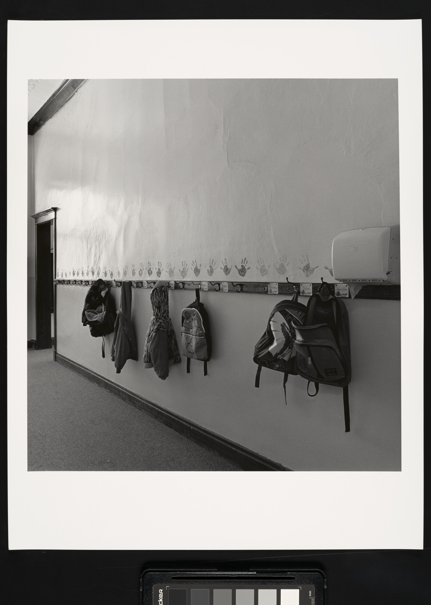 A black-and-white photograph of a hallway with kids&#039; backpacks and jackets hanging on hooks.