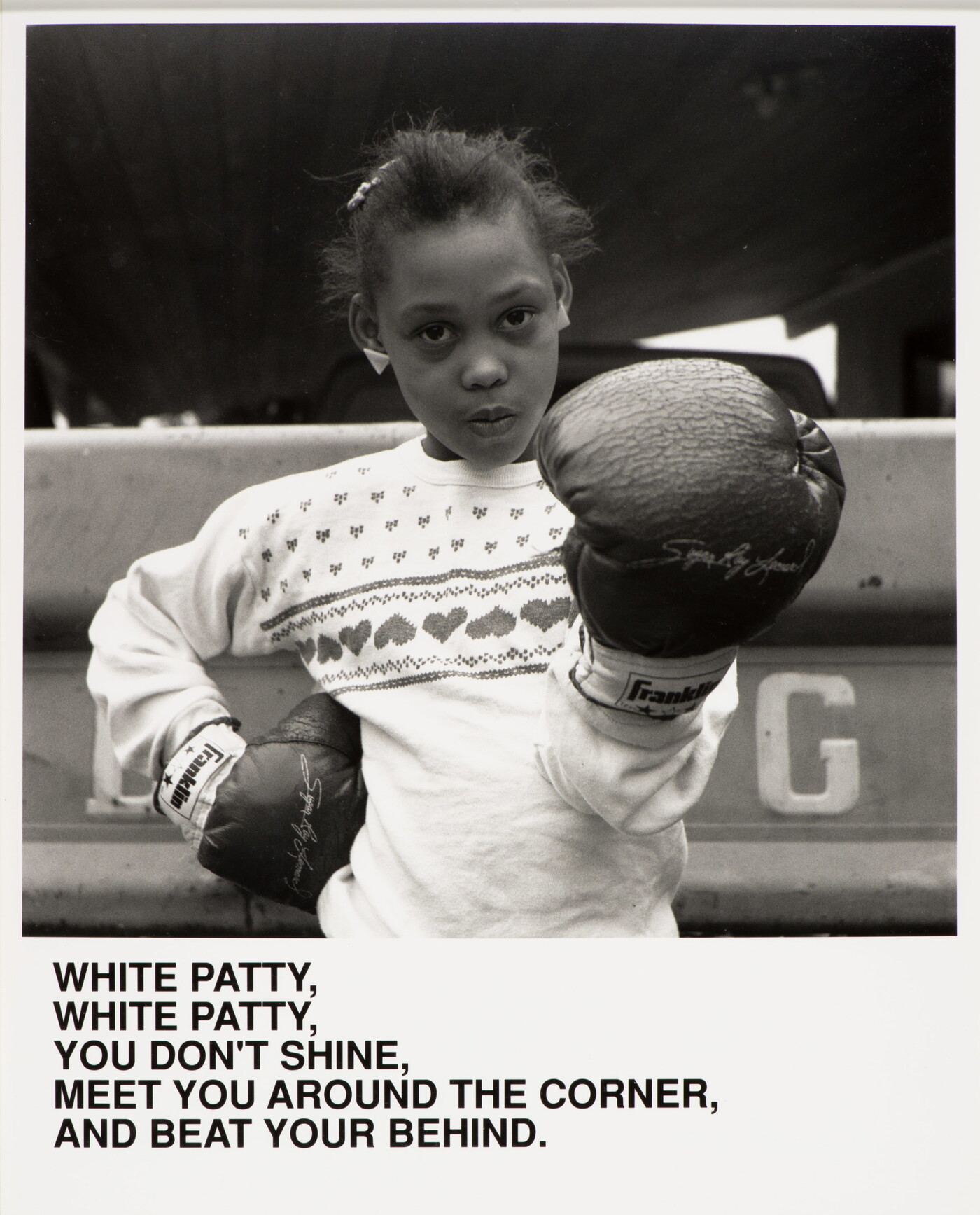 A black-and-white photograph of a Black girl wearing boxing gloves, one hand on her hip the other aimed at the viewer, above text that reads, &quot;White Patty, White Patty, You don&#039;t shine. Meet you around the corner and beat your behind.&quot;