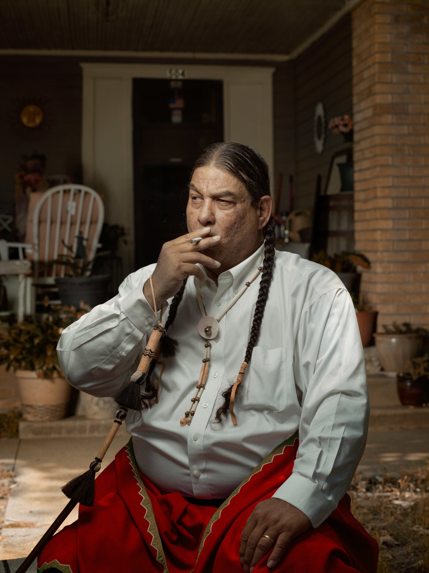 A color photograph of a seated Native American man with two, long braids smoking a cigarette in front of a porch.