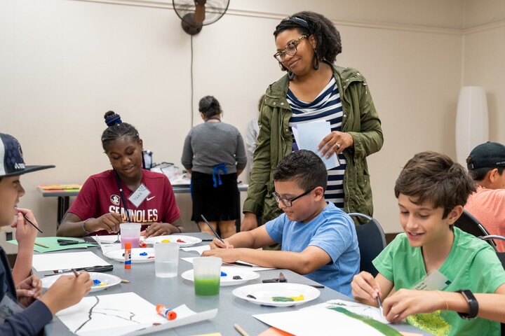 A standing woman looks at seated children doing an activity at a table.