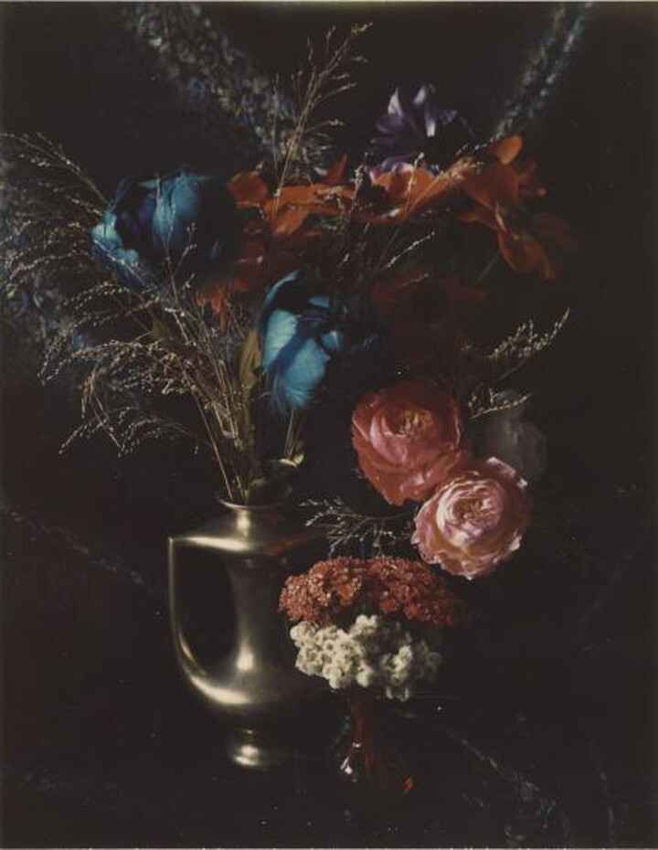 A color photograph of a variety of blue, purple, red, and pink flowers in a gold amphora-style vase.