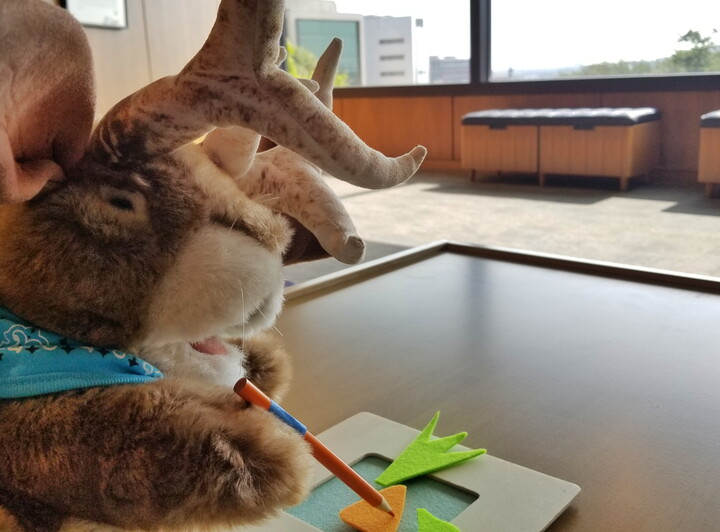 A jackalope hand puppet makes art at a table.