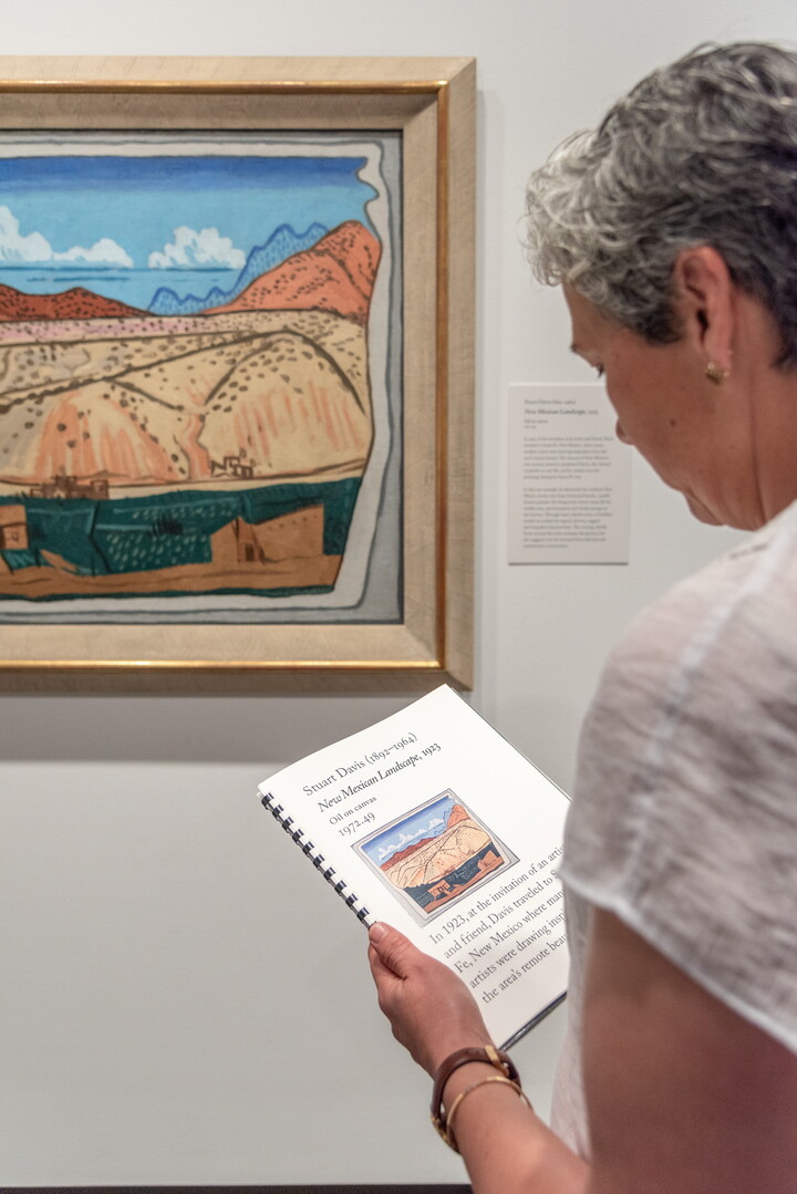 A person holds a large-print booklet as they stand in front of a painting.