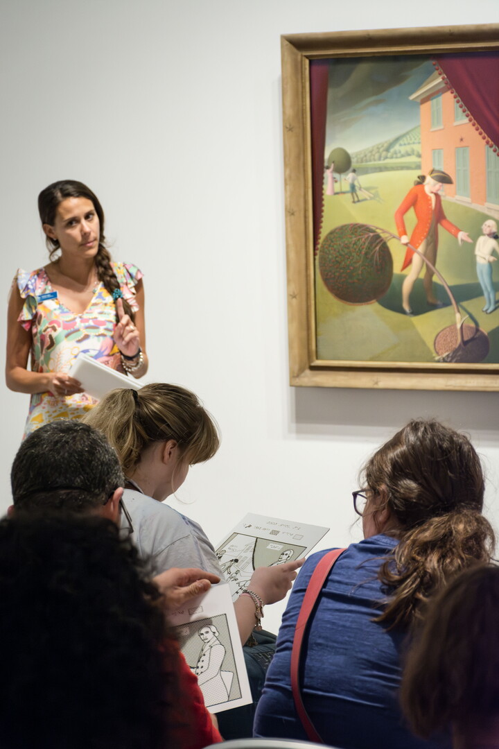 A White woman stands next to an artwork and in front of a group of adults.