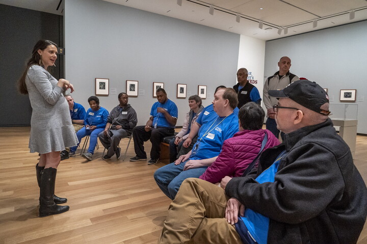A White woman stands in a Carter gallery of photographs while talking to a group of seated adults. 