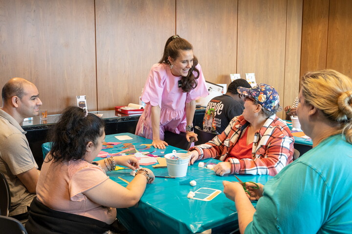 A White woman talks to adults seated around a table making art.