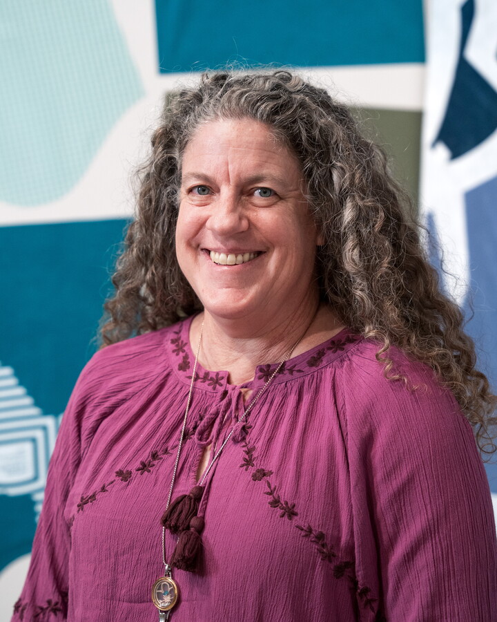 A headshot of a smiling White woman with long, curly salt-and-pepper hair in a gallery with cut clothing on the walls.