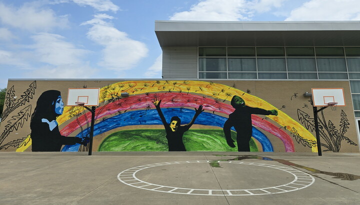 A colorful mural on the side of a brick building shows children playing and blowing dandelion "snow" on a rainbow backdrop.