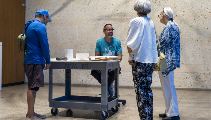 A middle-aged White man sits next to a rolling cart talking to visitors in a Carter gallery.