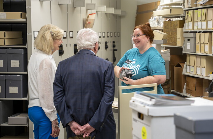 A smiling young White woman with red hair shows an object to a couple standing in a room lined with shelves..