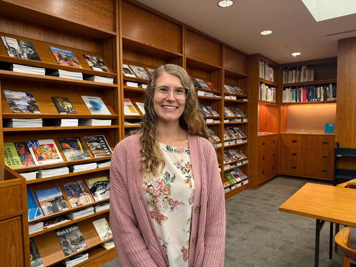 A smiling young White woman with long blonde hair and glasses poses in front of bookshelves in the Carter Library.